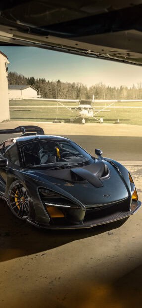 A McLaren Senna is parked inside a hangar with a small airplane visible outside on the runway, HD Phone Wallpaper