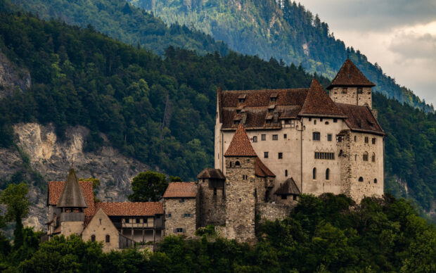 A historic stone castle stands surrounded by lush green forests in Liechtenstein, HD Desktop Wallpaper