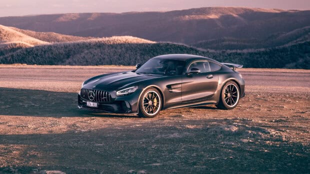 A black Mercedes Benz AMG GT is parked on a mountain road with hills in the background at sunset, 2K Desktop Wallpaper