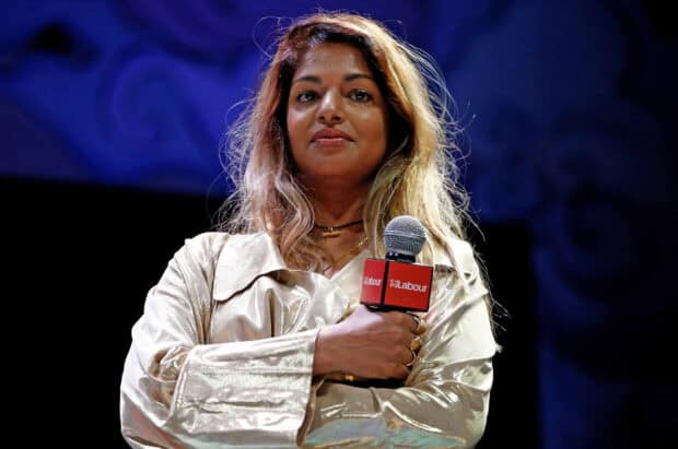 Mia rapper speaking seriously while holding a microphone with Labour logo, 2K Desktop Wallpaper