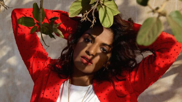 Mia rapper wears a red jacket under leafy shadows looking at the camera, HD Desktop Wallpaper
