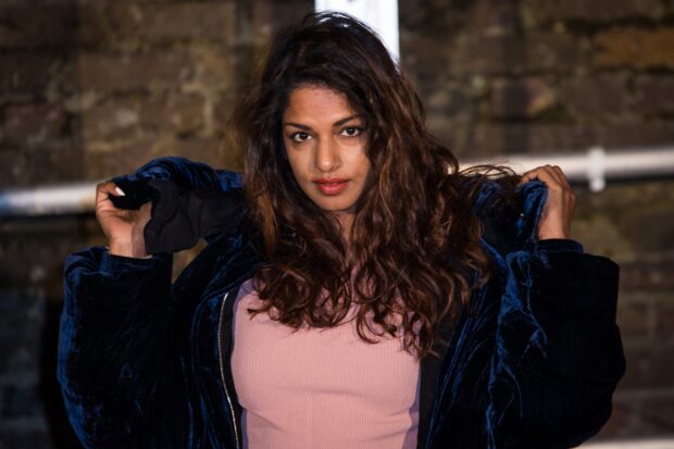 Mia rapper in a dark blue jacket posing confidently against a brick wall, 2K Desktop Wallpaper
