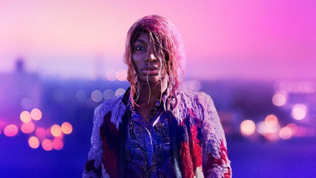 Michaela Coel stands against a vibrant cityscape with purple and pink hues at dusk, 2K Desktop Wallpaper
