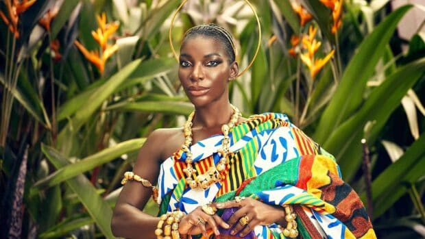 Michaela Coel wears colorful traditional attire and gold jewelry surrounded by lush greenery, HD Desktop Wallpaper