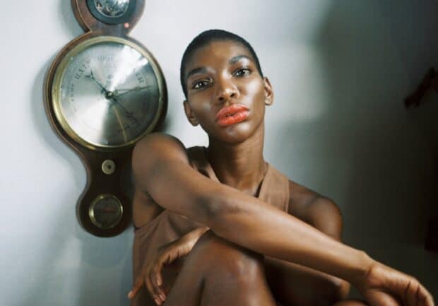 A portrait of Michaela Coel sitting with a calm expression near a wall clock, 2K Desktop Wallpaper