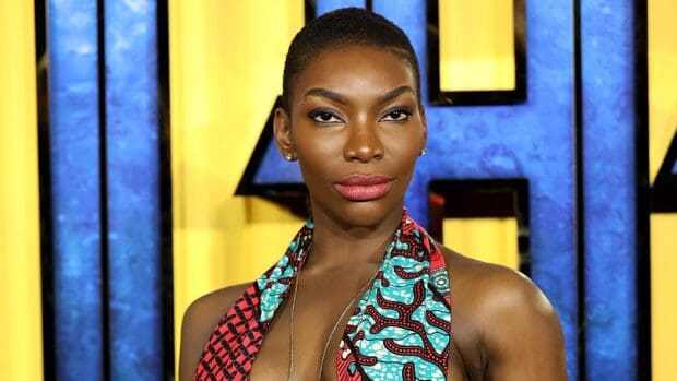 Michaela Coel wallpapers shows a confident woman with bright patterned clothing standing before a blue and yellow backdrop, 2K Desktop Wallpaper