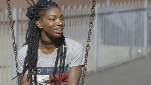 Michaela Coel smiles while sitting on a swing in an outdoor area, HD Desktop Wallpaper