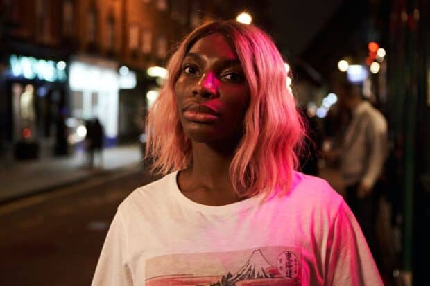 Michaela Coel stands on a city street illuminated by pink light in this portrait, HD Desktop Wallpaper