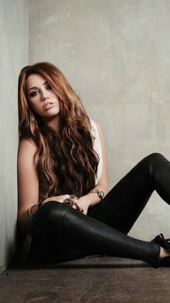 Miley Cirus wallpapers feature her sitting casually against a wall with long highlighted hair and stylish black pants, 2K Mobile Wallpaper