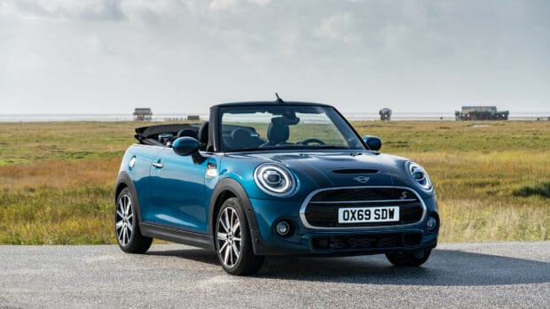 A blue Mini Cooper convertible is parked near a grassy field under a cloudy sky, 2K Desktop Wallpaper