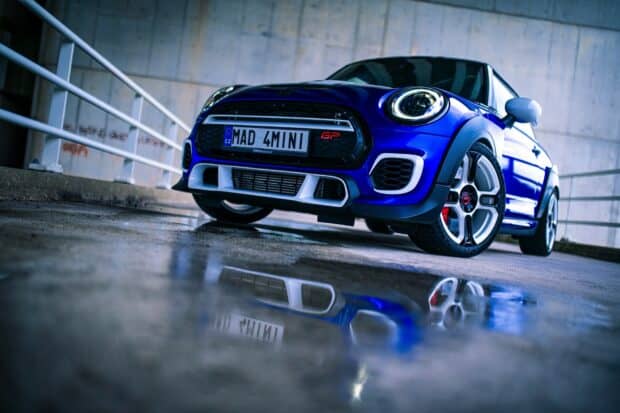 A blue Mini Cooper with distinctive headlights is parked near a concrete wall reflecting on the wet ground, HD Desktop Wallpaper