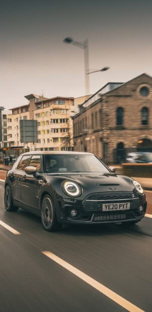 A black Mini Cooper is driving quickly on a city street with buildings in the background, 2K Mobile Wallpaper