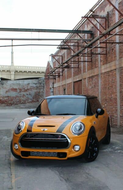 A bright yellow Mini Cooper is parked near old brick walls with metal beams above, HD Mobile Wallpaper