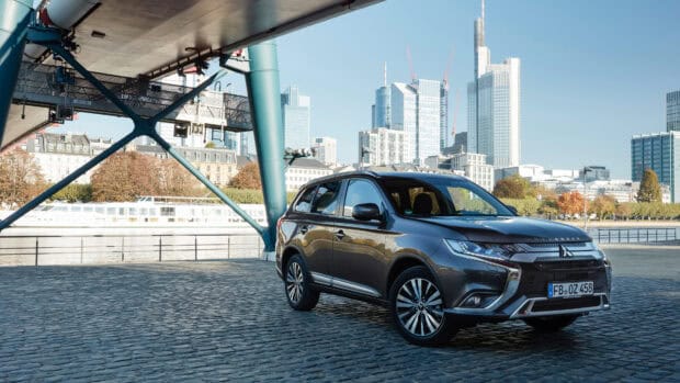 A Mitsubishi Outlander is parked on a cobblestone surface near a river with city buildings in the background, 4K Desktop Wallpaper