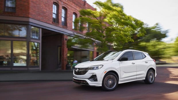 A white Buick Encore drives quickly past a brick building with blurred green trees in the background, 4K Desktop Wallpaper