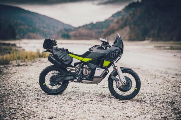 A modern Enduro motorbike is parked on rocky ground by a river with hills in the background