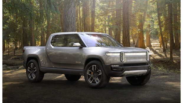 A silver Rivian electric truck is parked in a sunlit forest with tall trees surrounding it, HD Desktop Wallpaper