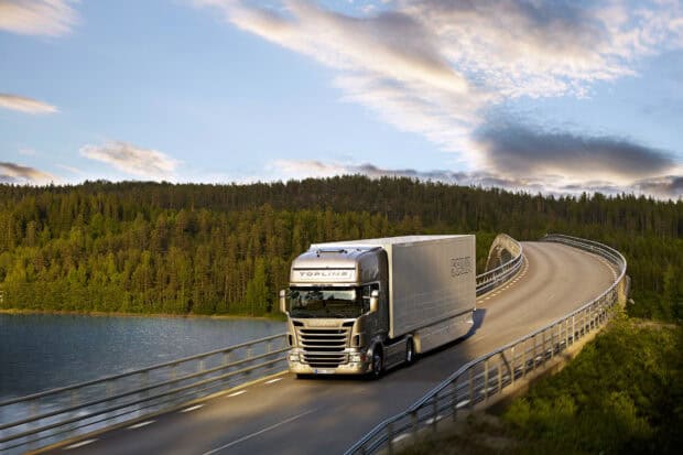 A silver Scania truck drives along a raised curved bridge surrounded by forest and water under a blue sky, HD Desktop Wallpaper