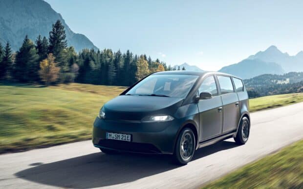 A Sono Motors vehicle drives along a rural mountain road with trees and peaks in the background, 2K Desktop Wallpaper