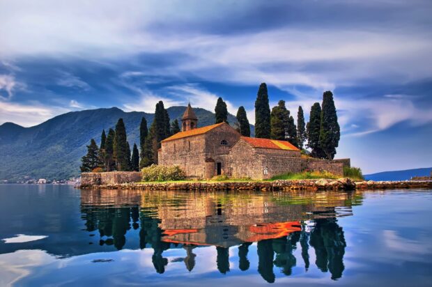 A small island with a church and cypress trees is reflected in calm water in Montenegro, 2K Desktop Wallpaper