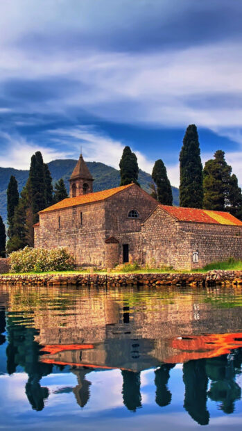 A historic stone church reflects on the calm water near Montenegro with tall trees and mountains, HD Phone Wallpaper
