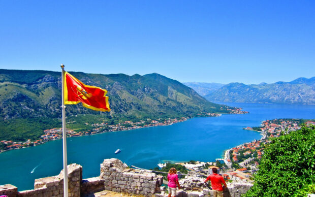 A scenic view of Montenegro with a red flag overlooking the blue bay and mountains, 2K Desktop Wallpaper
