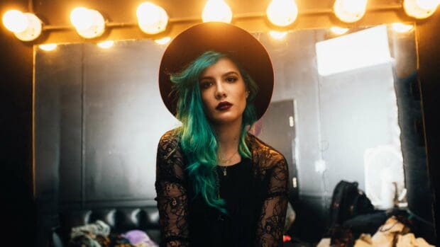 A moody portrait of Halsey with teal hair and dark lipstick sitting in a lit dressing room