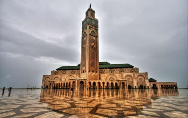 The Hassan II Mosque stands tall with detailed designs reflecting on wet courtyard, 2K Desktop Wallpaper
