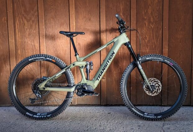 A green mountain Transition bike stands against a wooden wall with visible suspension and tires, HD Desktop Wallpaper