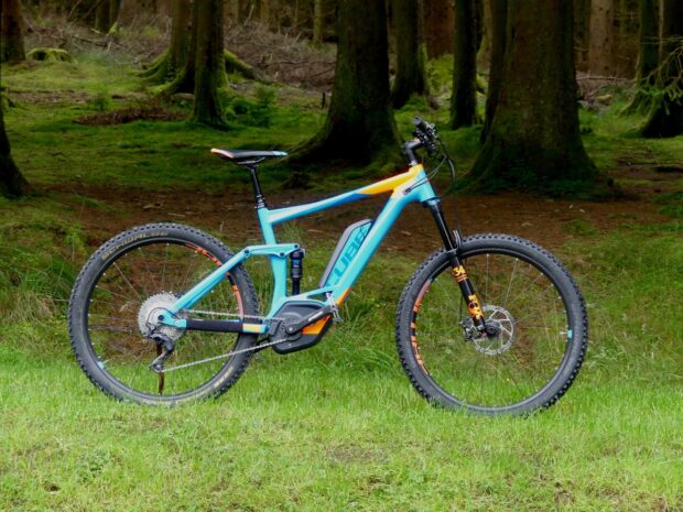 A blue and orange mountain bike stands on grass near trees featuring Cube Bikes Wallpapers branding, HD Desktop Wallpaper