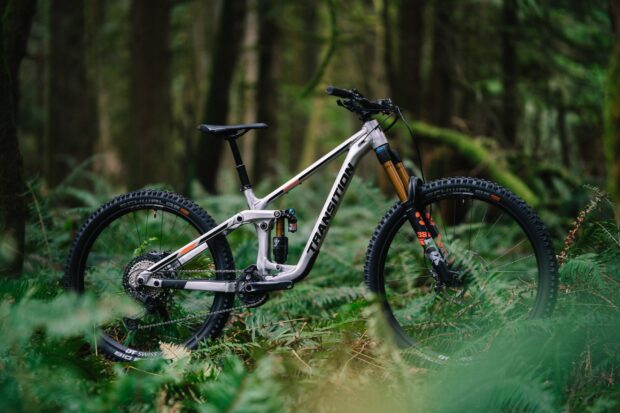 A silver mountain bike with Transition Bikes logo is resting on a grassy forest floor surrounded by trees, 2K Desktop Wallpaper