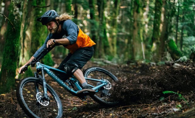 A mountain biker rides a Transition bike on a muddy forest trail, kicking up dirt and leaves, HD Desktop Wallpaper