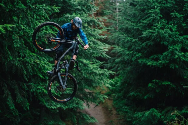 A mountain biker is doing a trick on a trail surrounded by green trees in the forest, HD Desktop Wallpaper