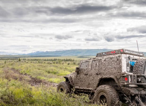 A mud covered off road vehicle is driving through a grassy and muddy terrain with mountains in the background, HD Desktop Wallpaper