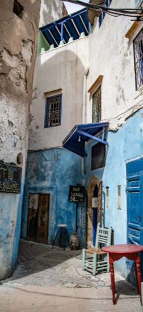 A narrow Moroccan alley features blue walls with rustic doors and a small table under natural light, 2K Phone Wallpaper