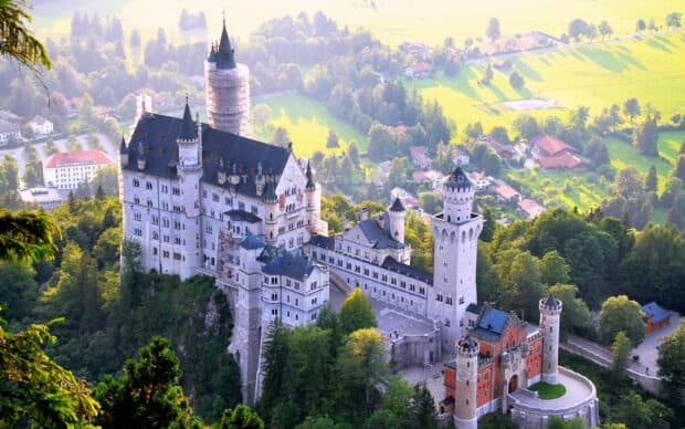 Neuschwanstein Castle stands prominently surrounded by trees and green fields on a sunny day, 2K Desktop Wallpaper