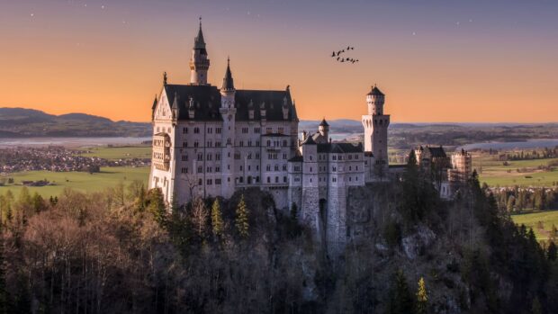 Neuschwanstein Castle stands majestically on a hill with a sunset sky and birds flying, 4K Desktop Wallpaper