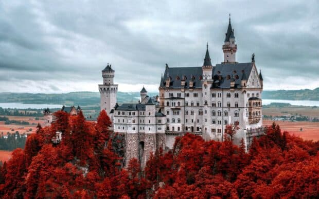 Neuschwanstein Castle stands surrounded by red autumn trees under a cloudy sky, HD Desktop Wallpaper