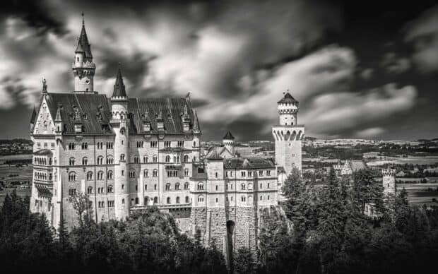 A dramatic black and white photo of Neuschwanstein Castle surrounded by trees and hills, 2K Desktop Wallpaper
