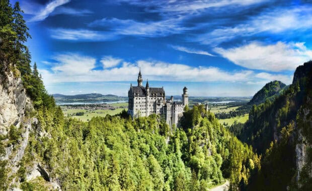 The Neuschwanstein Castle stands surrounded by lush green forest under a partly cloudy blue sky, 2K Desktop Wallpaper