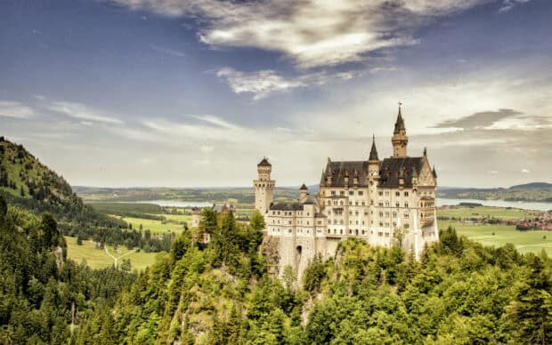 A scenic view of Neuschwanstein Castle surrounded by forest and fields, 2K Desktop Wallpaper