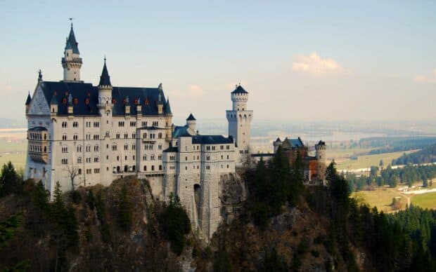 Neuschwanstein Castle stands on a rugged hill surrounded by trees and a vast landscape, 2K Desktop Wallpaper