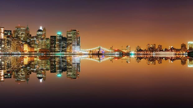 The New York skyline shows beautifully lit buildings and a bridge reflected on calm water at night, 2K Desktop Wallpaper