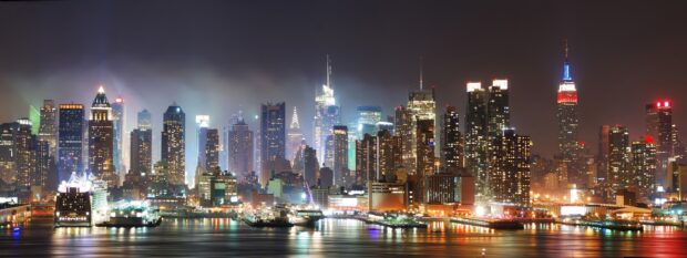 The New York skyline at night shows illuminated skyscrapers over the water, 2K Desktop Wallpaper