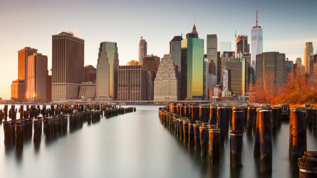The New York skyline features iconic buildings glowing at sunset with wooden posts in the water, 4K Desktop Wallpaper