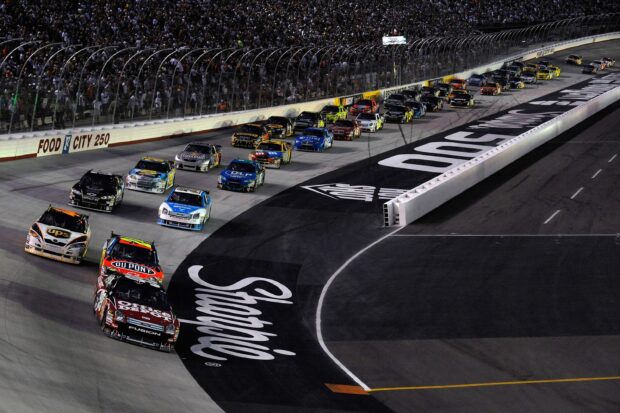A group of race cars speeding along a well lit race track with fans in the stands, 2K Desktop Wallpaper