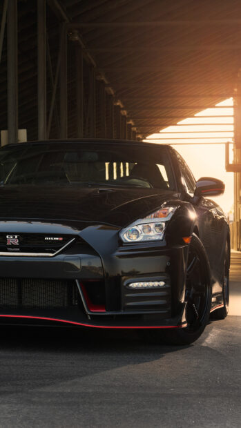 A black Nissan GT R is parked under a structure with sunlight glowing behind it, 4K Phone Wallpaper