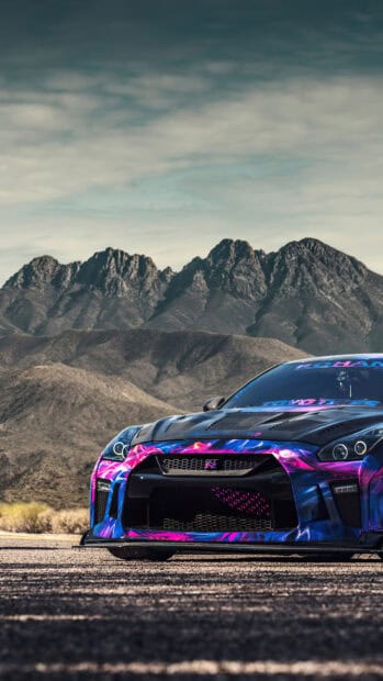 A Nissan GT R with vibrant blue and pink pink flames design is parked on a road in front of rocky mountains, 4K Phone Wallpaper