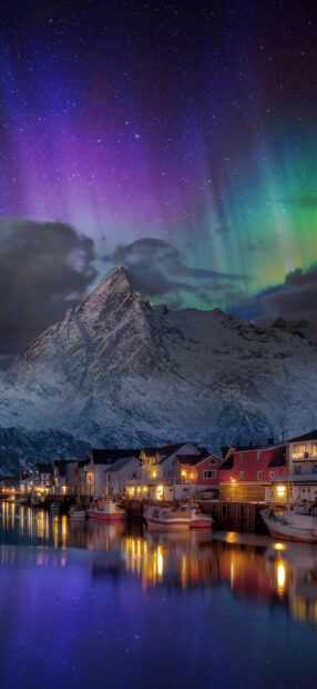 The Norway Wallpapers shows vibrant northern lights illuminating a snowy mountain and colorful village by the water at night, 2K Phone Wallpaper