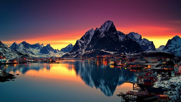 A vibrant sunset reflects over the Norwegian fjord surrounded by snow capped mountains and village lights, 4K Desktop Wallpaper